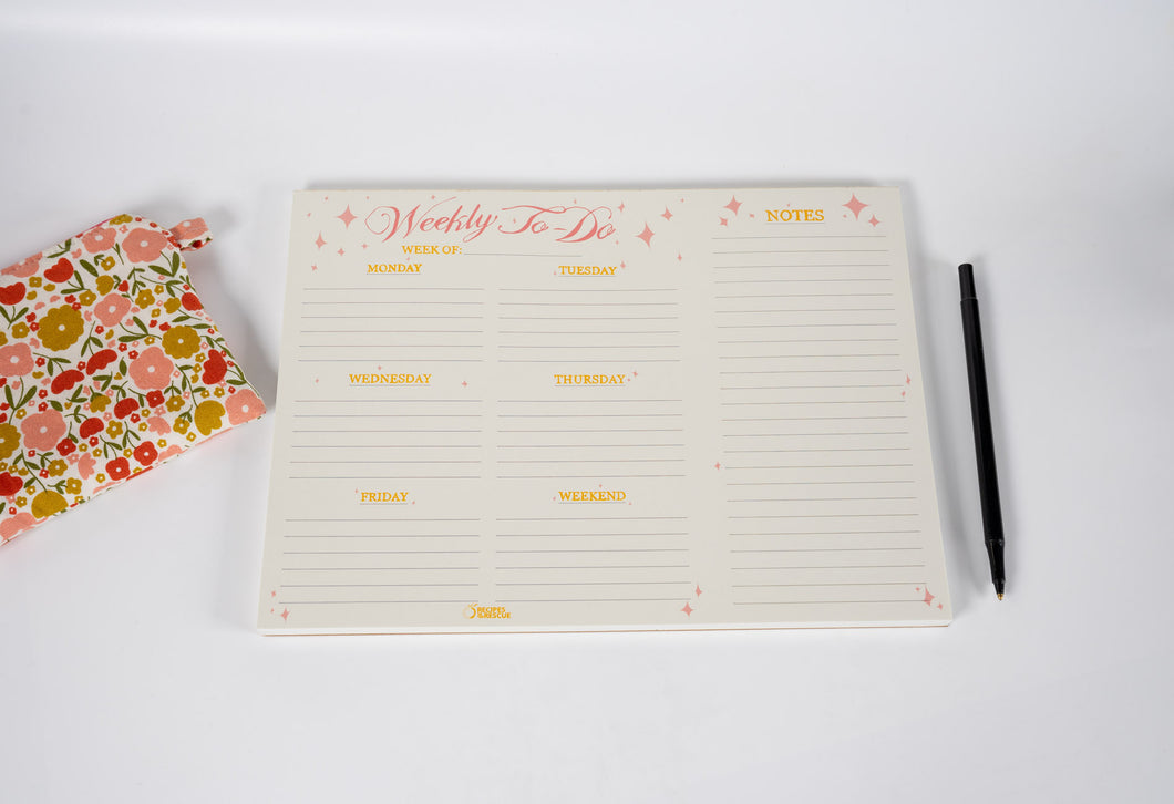 Weekly to-do list pad with a pen and floral notebook on a white background